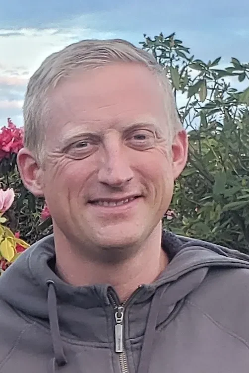 Man with short blond hair smiling at the camera, wearing a gray zip-up hoodie, photographed outdoors in a garden with pink and red flowering shrubs and green foliage against a cloudy sky