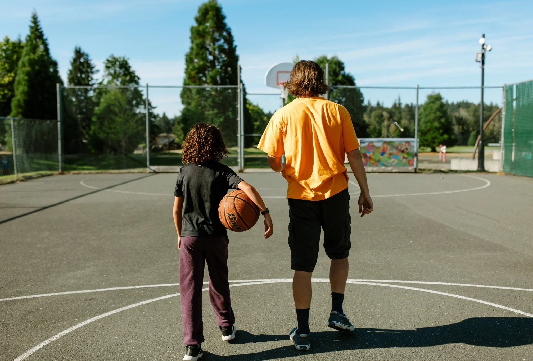 KidStart-playing-basketball An adult and a child walk onto an outdoor basketball court, the child carrying a basketball.