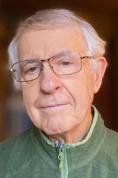 Portrait of an older man wearing glasses and a green zip-up fleece, softly lit with a warm, blurred background, conveying calm, thoughtfulness, and approachability.