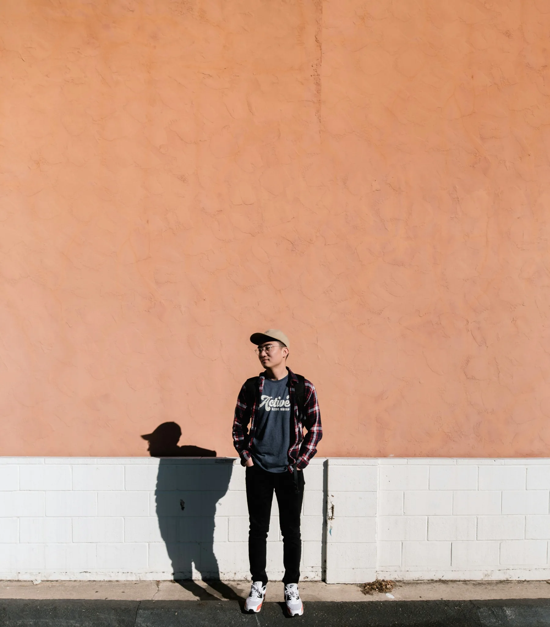 boy-standing-against-wall Young man standing against a peach-colored wall wearing casual streetwear, minimalist urban portrait with strong sunlight and shadow.