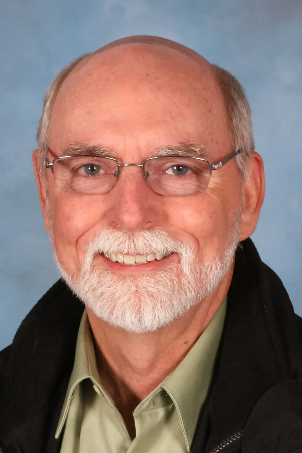 Portrait of an older man with glasses and a white beard smiling against a light blue studio background, conveying warmth, approachability, and professionalism.
