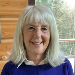 Smiling woman with shoulder-length silver-gray hair and bangs, wearing a royal blue sweater over a white collared shirt, photographed indoors near a window with a wooden wall and balcony railing with trees visible in the background.