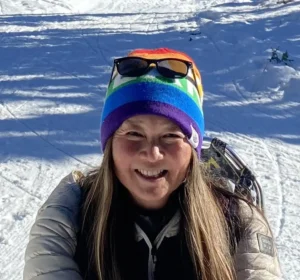 Woman with long brown hair smiling brightly in a snowy ski setting, wearing a colorful rainbow-striped beanie with sunglasses resting on top and a tan quilted jacket, with snow-covered slopes visible in the background.