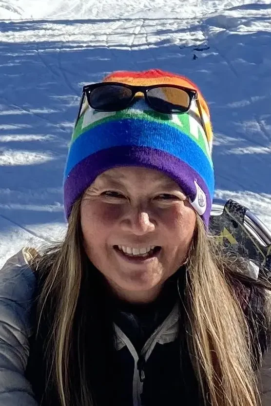 Woman with long brown hair smiling brightly in a snowy ski setting, wearing a colorful rainbow-striped beanie with sunglasses resting on top and a tan quilted jacket, with snow-covered slopes visible in the background.