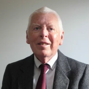 Professional headshot of an older man with white hair and blue eyes, smiling warmly at the camera, wearing a gray herringbone tweed blazer, white dress shirt, and burgundy polka dot tie against a plain light gray background.