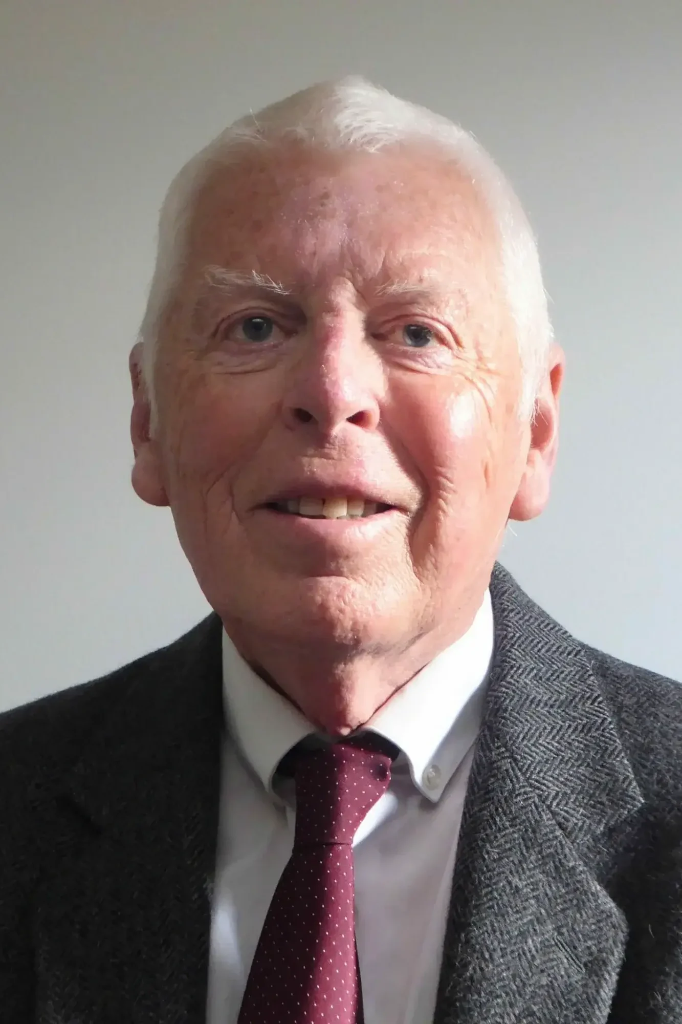 Professional headshot of an older man with white hair and blue eyes, smiling warmly at the camera, wearing a gray herringbone tweed blazer, white dress shirt, and burgundy polka dot tie against a plain light gray background.