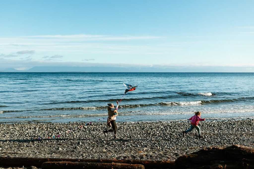 Two children run along a rocky beach shoreline on a sunny day, flying a colourful kite. One child holds the kite aloft while the other runs alongside with arms outstretched. Calm ocean waves and a distant mountain range are visible in the background under a pale blue sky.