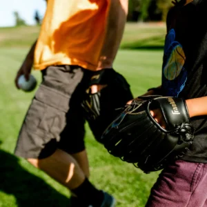 An adult and a child walking across a sunny green field, one wearing an orange shirt and grey shorts holding a baseball, the other in a dark graphic t-shirt and maroon pants carrying a black Franklin baseball glove.