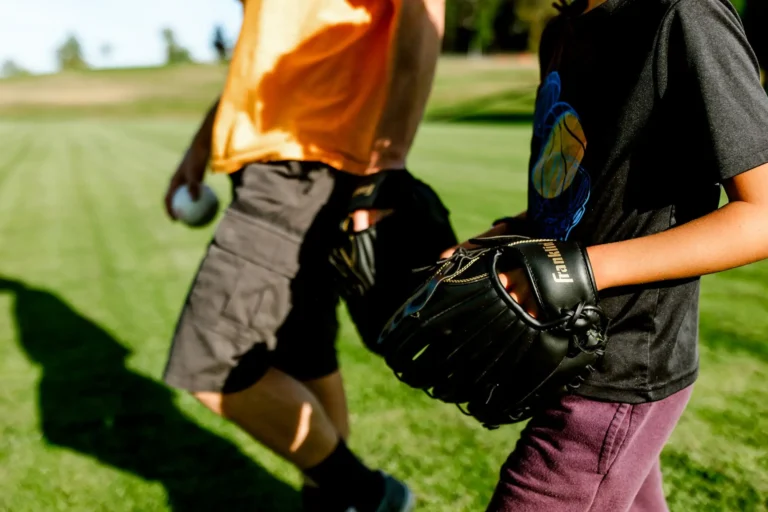 An adult and a child walking across a sunny green field, one wearing an orange shirt and grey shorts holding a baseball, the other in a dark graphic t-shirt and maroon pants carrying a black Franklin baseball glove.