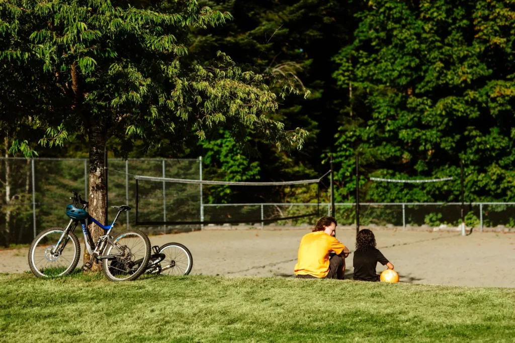 An adult in an orange shirt and a child sit side by side on the edge of a sandy sports court in a park, holding a ball and talking, with two bicycles leaned against a tree nearby and lush green trees in the background.